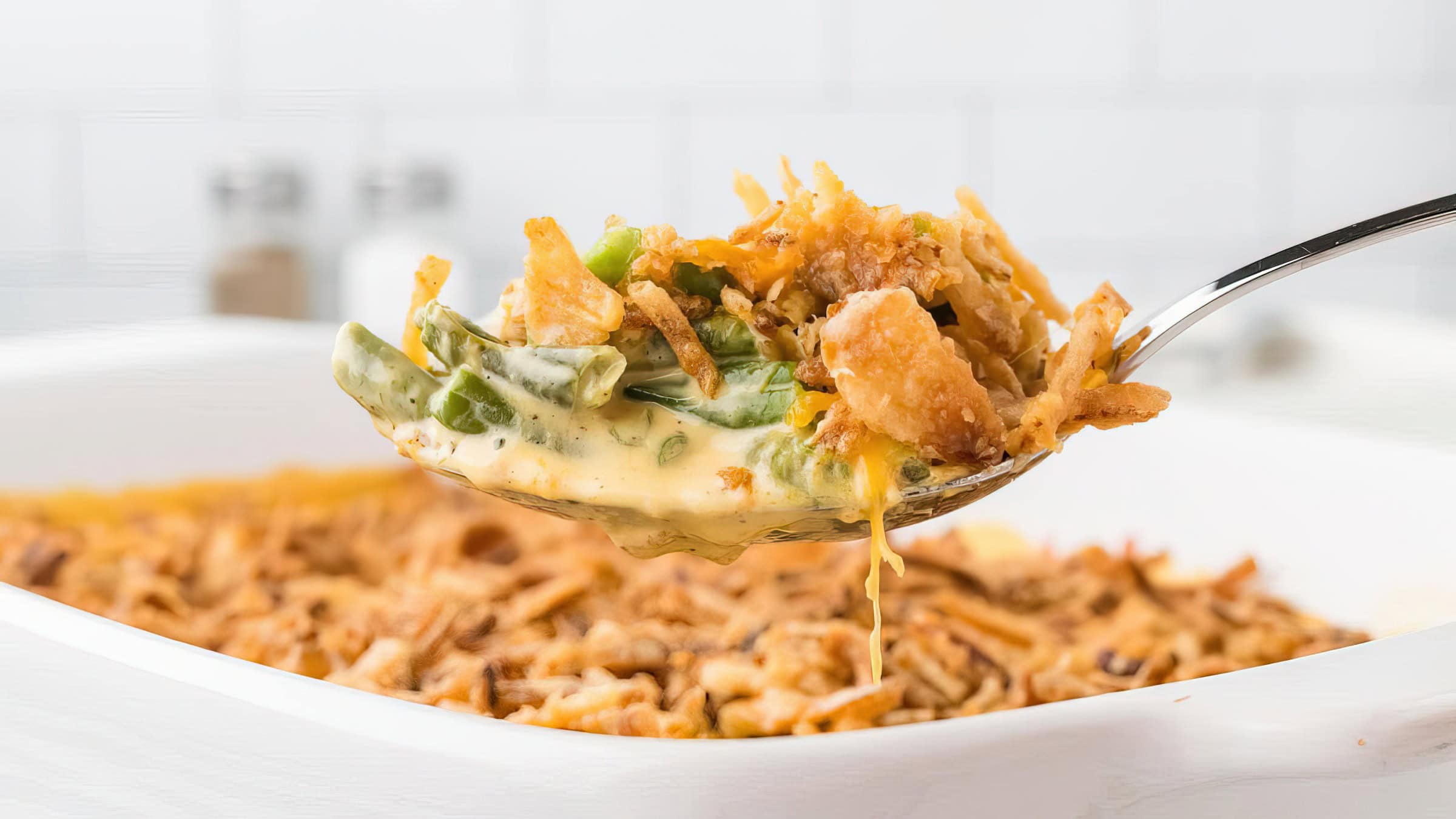 A spoonful of green bean casserole is lifted above a white dish. The casserole features green beans, creamy sauce, and a crunchy topping. The background is softly blurred, showing a white kitchen setting.