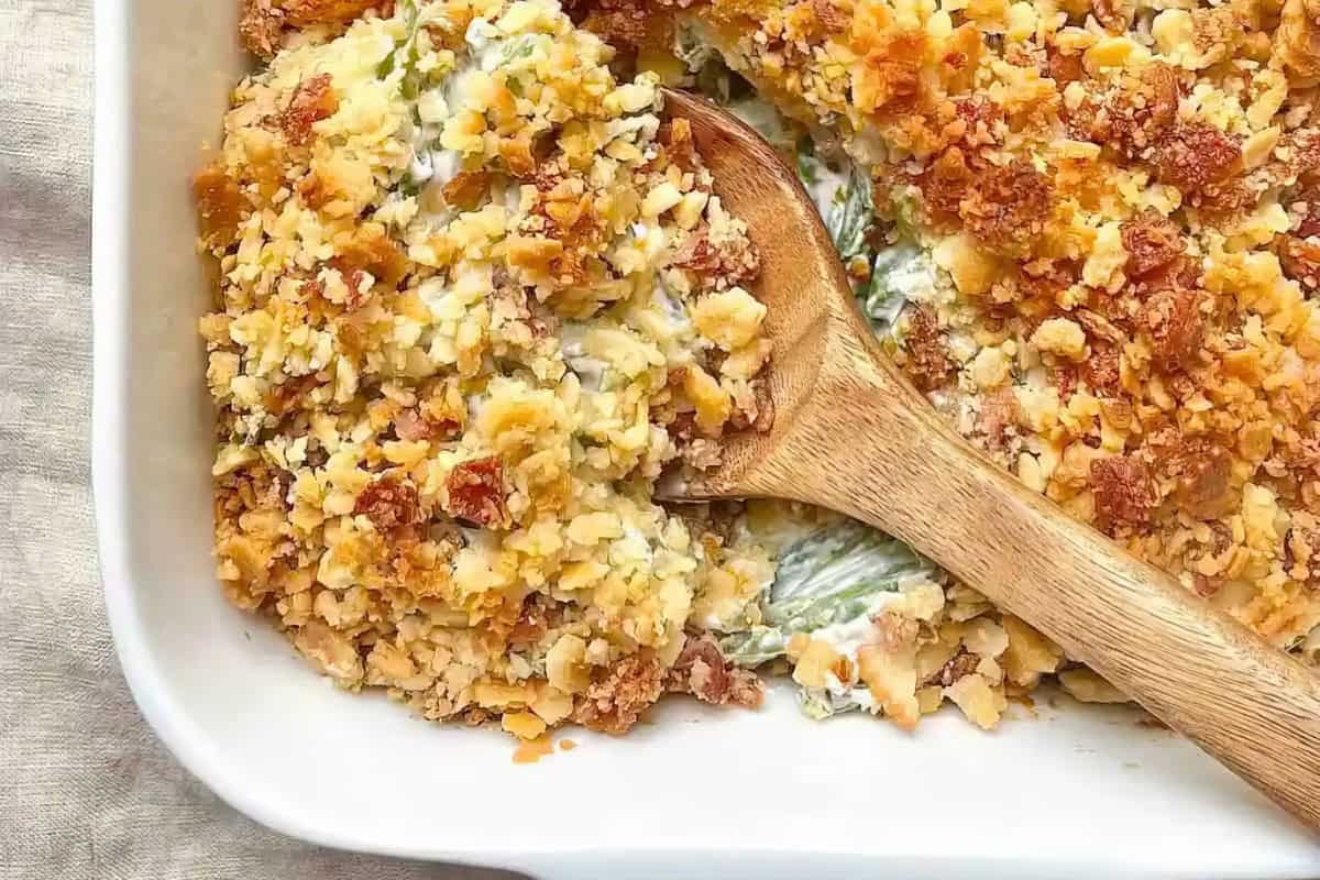 The image shows a casserole dish filled with a creamy mixture, topped with a golden, crumbly crust. A wooden spoon partially uncovers the filling.
