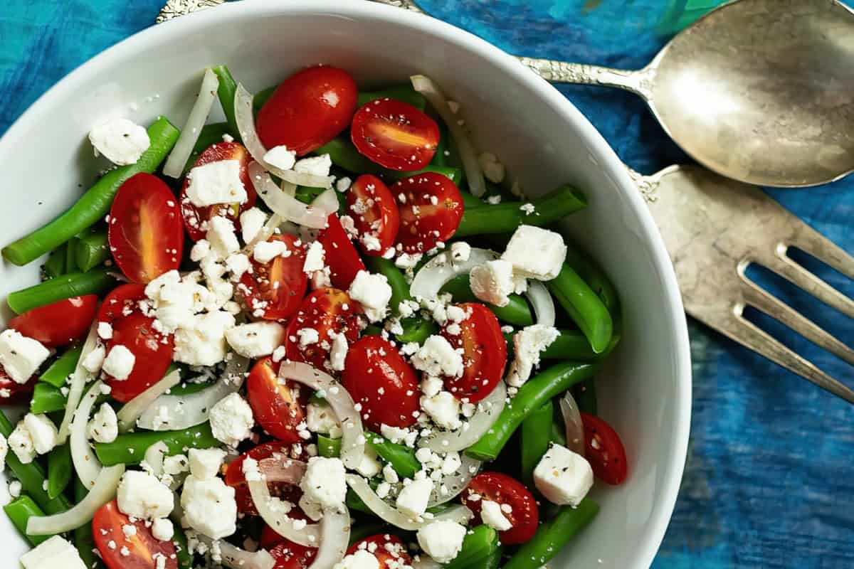 A salad bowl containing green beans, cherry tomatoes, onion slices, and crumbled feta cheese, set against a blue background, with a spoon and fork nearby.