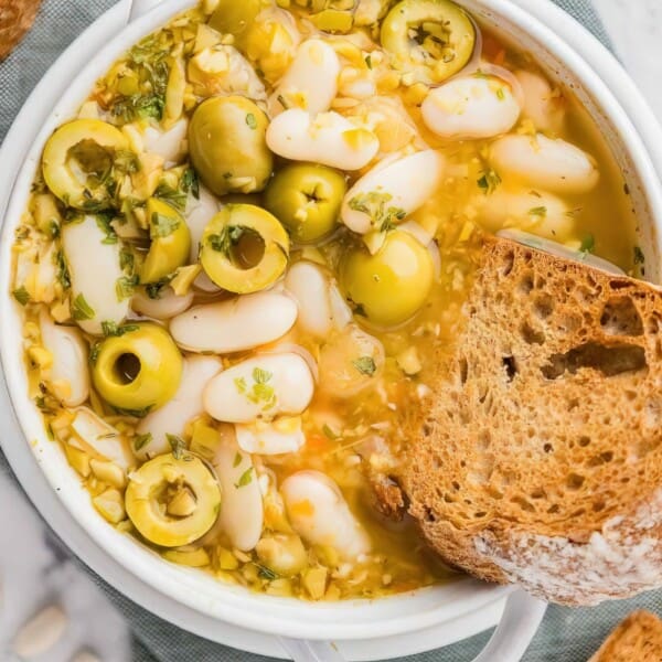 A bowl of soup with white beans, green olives, and herbs, accompanied by a slice of crusty bread on a marble surface.