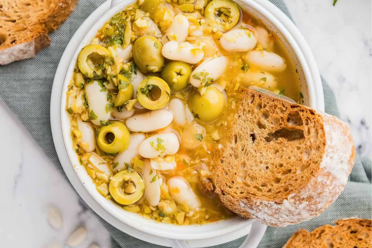 A bowl of soup with white beans, green olives, and herbs, accompanied by a slice of crusty bread on a marble surface.
