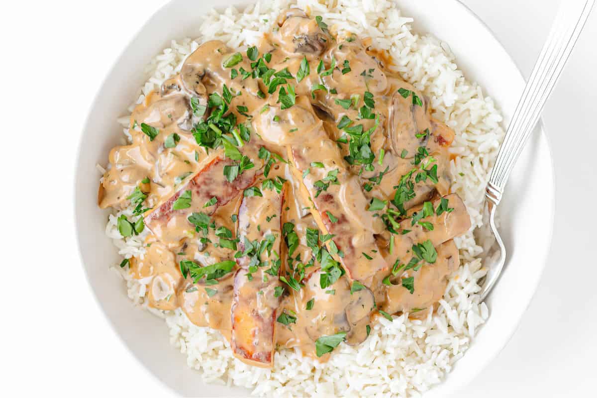 A bowl of white rice topped with creamy mushroom and tomato sauce, garnished with chopped parsley, with a fork resting on the side.
