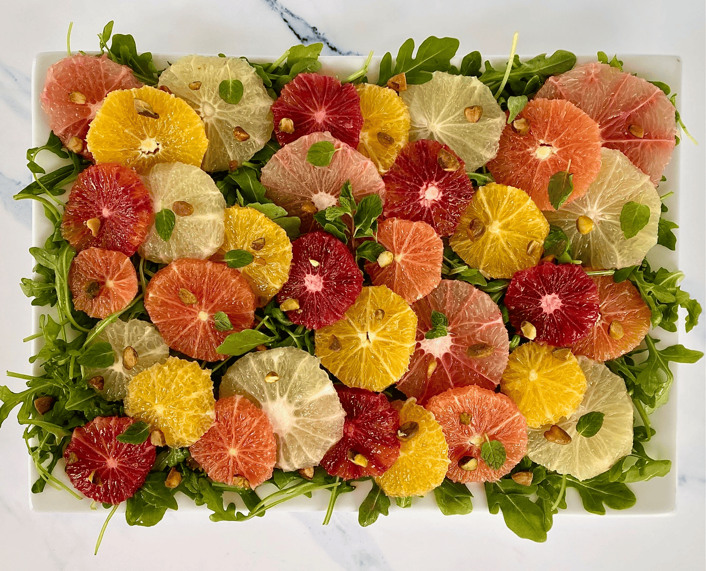 A colorful salad arranged on a rectangular platter features slices of various citrus fruits, including grapefruit, blood orange, and orange. The slices are laid over a bed of fresh arugula and garnished with pistachios and small green leaves.