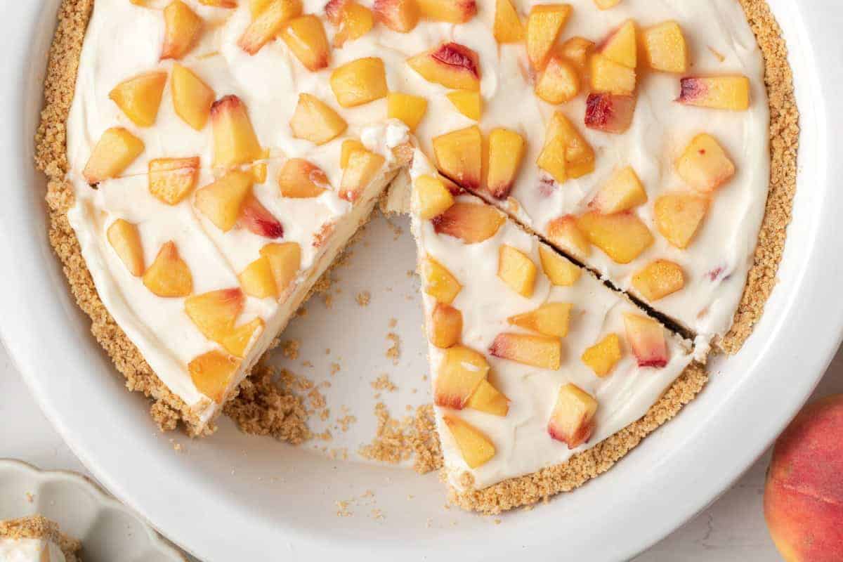 A fruit cream pie with a graham cracker crust and peach pieces on top, missing one slice, in a white pie dish.
