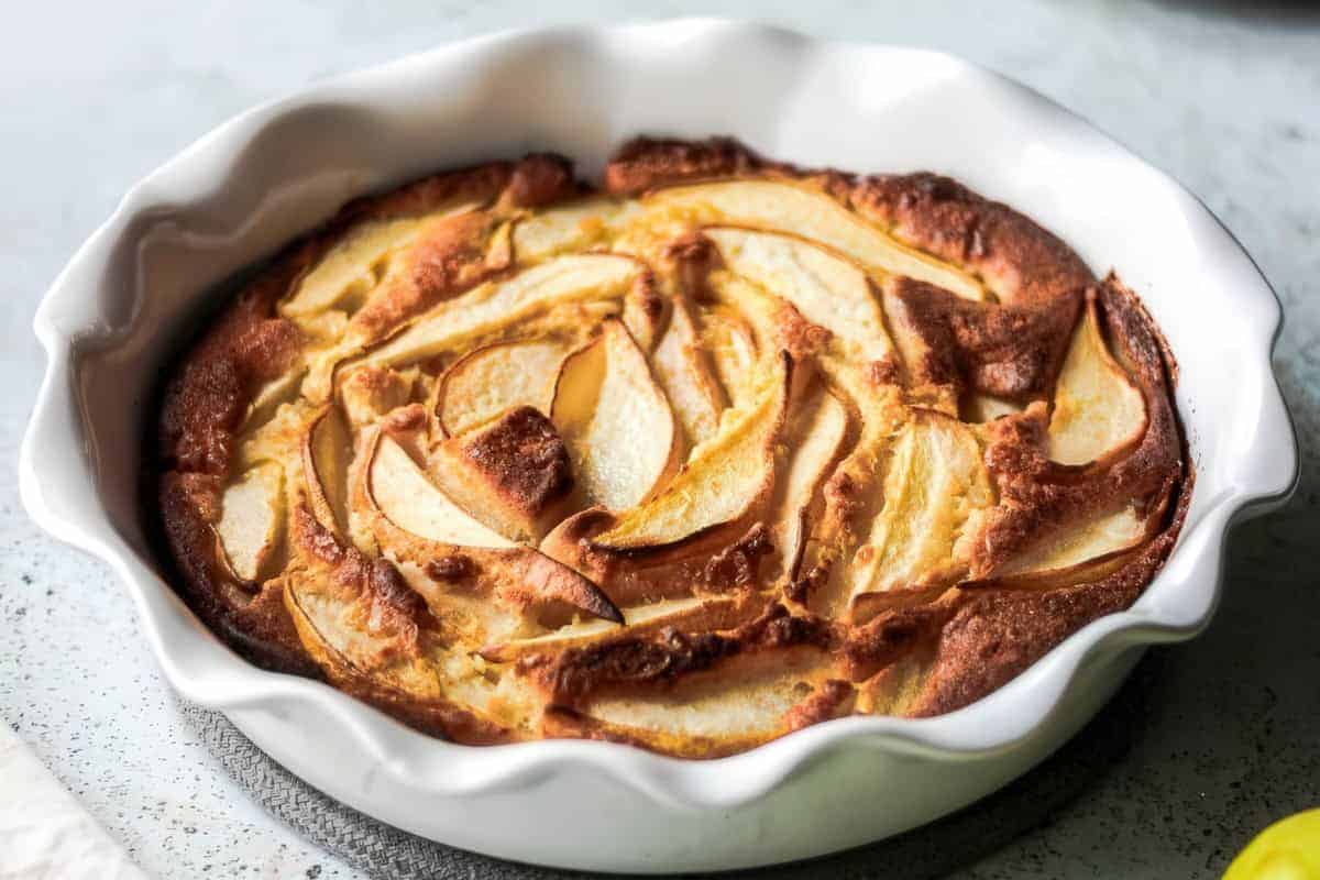 A baked dessert in a white fluted dish with golden-brown edges and layered apple slices arranged neatly on top.