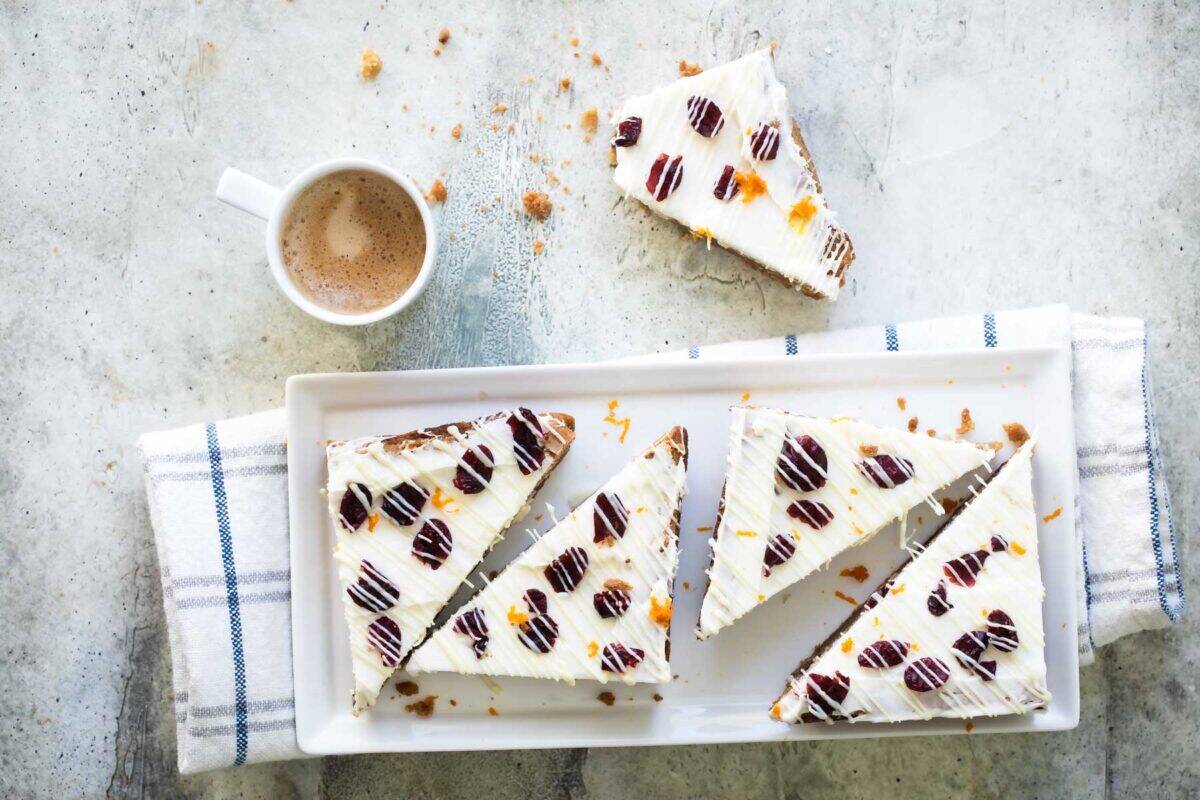 Rectangular white plate with five triangular cranberry-studded frosted desserts topped with chocolate pieces, beside a cup of coffee.