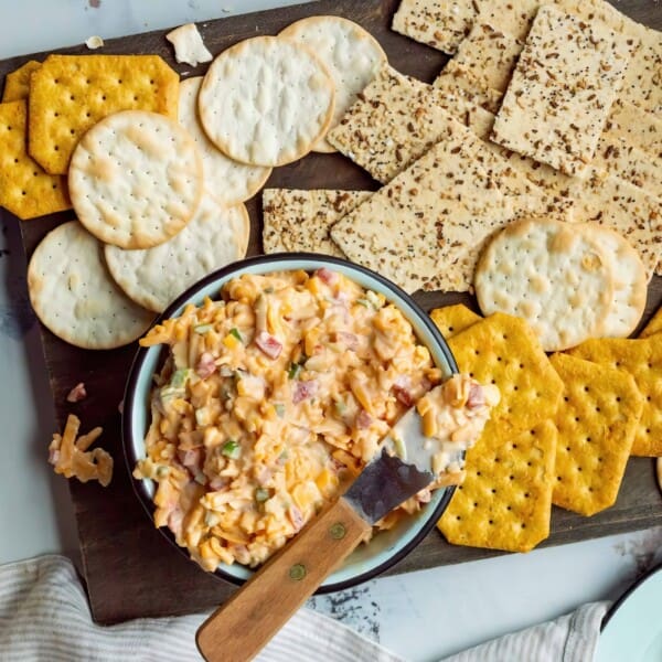 A bowl of pimento cheese with a spreader, surrounded by various crackers on a wooden board.