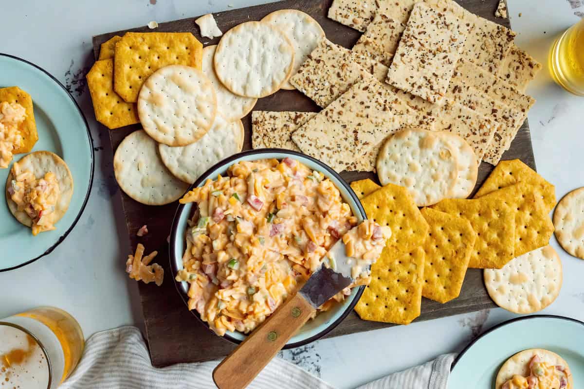 A bowl of pimento cheese with a spreader, surrounded by various crackers on a wooden board.