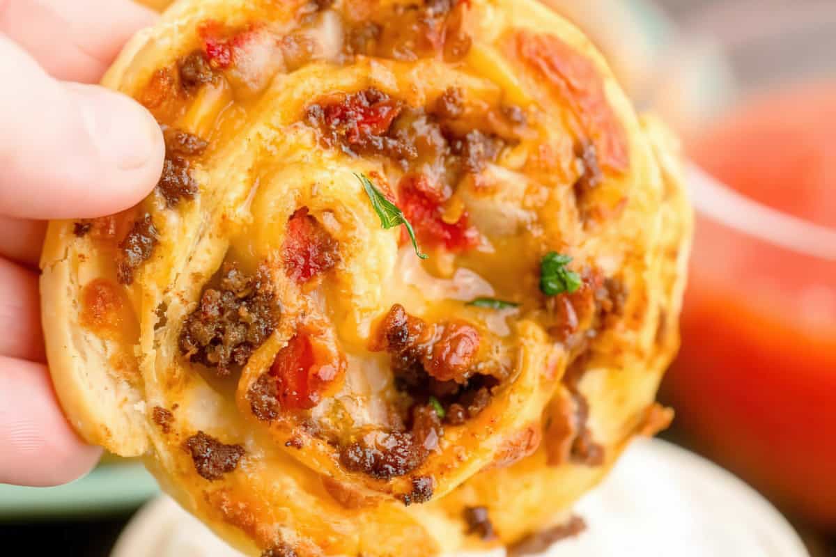 A close-up of a hand holding a pizza roll filled with cooked ground beef, melted cheese, and red bell peppers. The roll is golden brown with some herbs visible, and there's a blurred background with a red sauce.