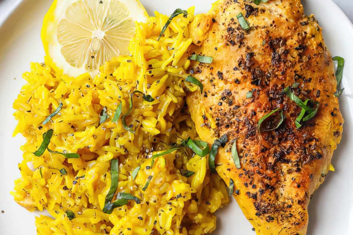 The image shows a plate with grilled chicken breast, yellow rice garnished with herbs, and a slice of lemon.