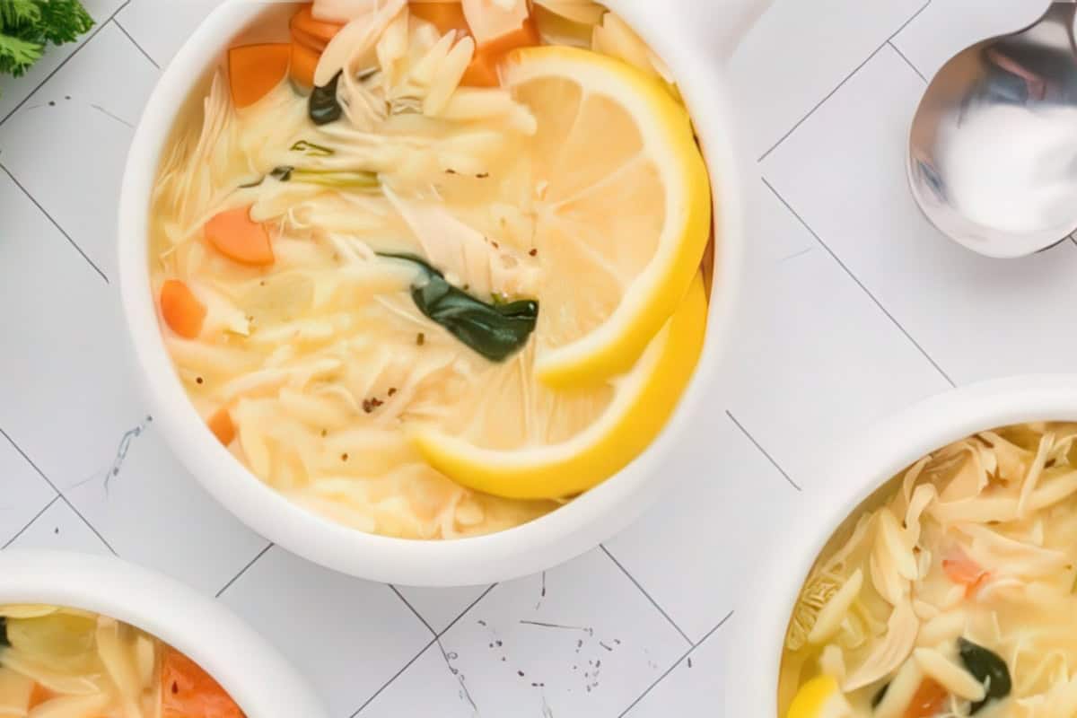 A bowl of soup with noodles, sliced lemons, carrots, and greens, placed on a grid-patterned surface. A spoon is nearby, and part of another bowl is visible at the bottom right corner.