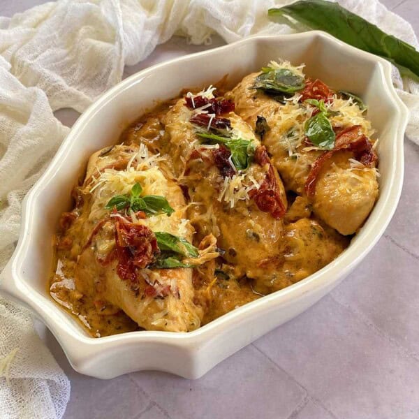 Baked chicken breasts with sun-dried tomatoes, melted cheese, and fresh basil in creamy sauce in a white oval dish.