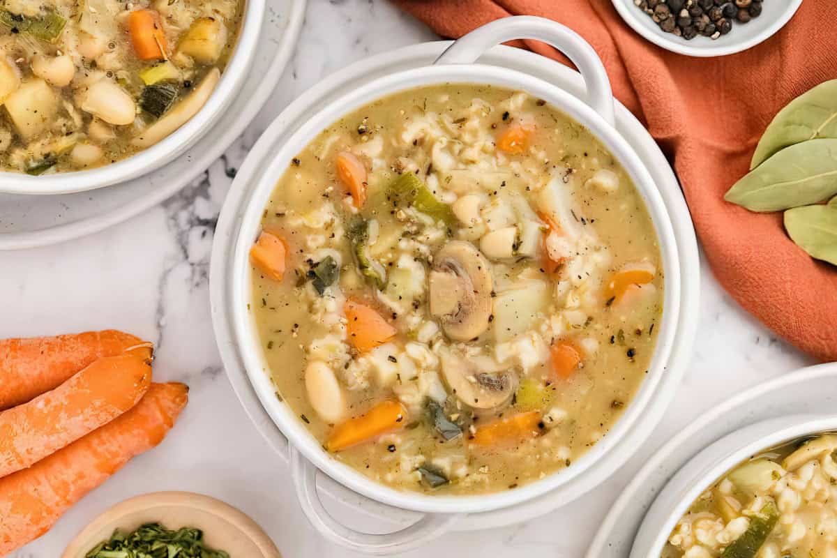 A bowl of vegetable and barley soup with carrots and mushrooms is placed on a marble surface, surrounded by whole carrots, bay leaves, and a bowl of peppercorns.