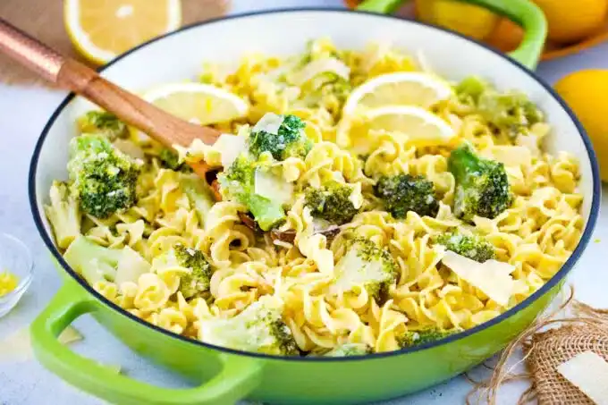 A green pan containing pasta, broccoli, lemon slices, and cheese shavings with a wooden spoon resting inside.