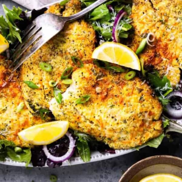 A plate of breaded baked fish fillets on mixed greens, garnished with lemon wedges and sliced green onions, with a fork on top.