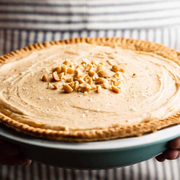A person holds a peanut butter pie topped with chopped peanuts, featuring a golden crust and creamy filling, on striped fabric.
