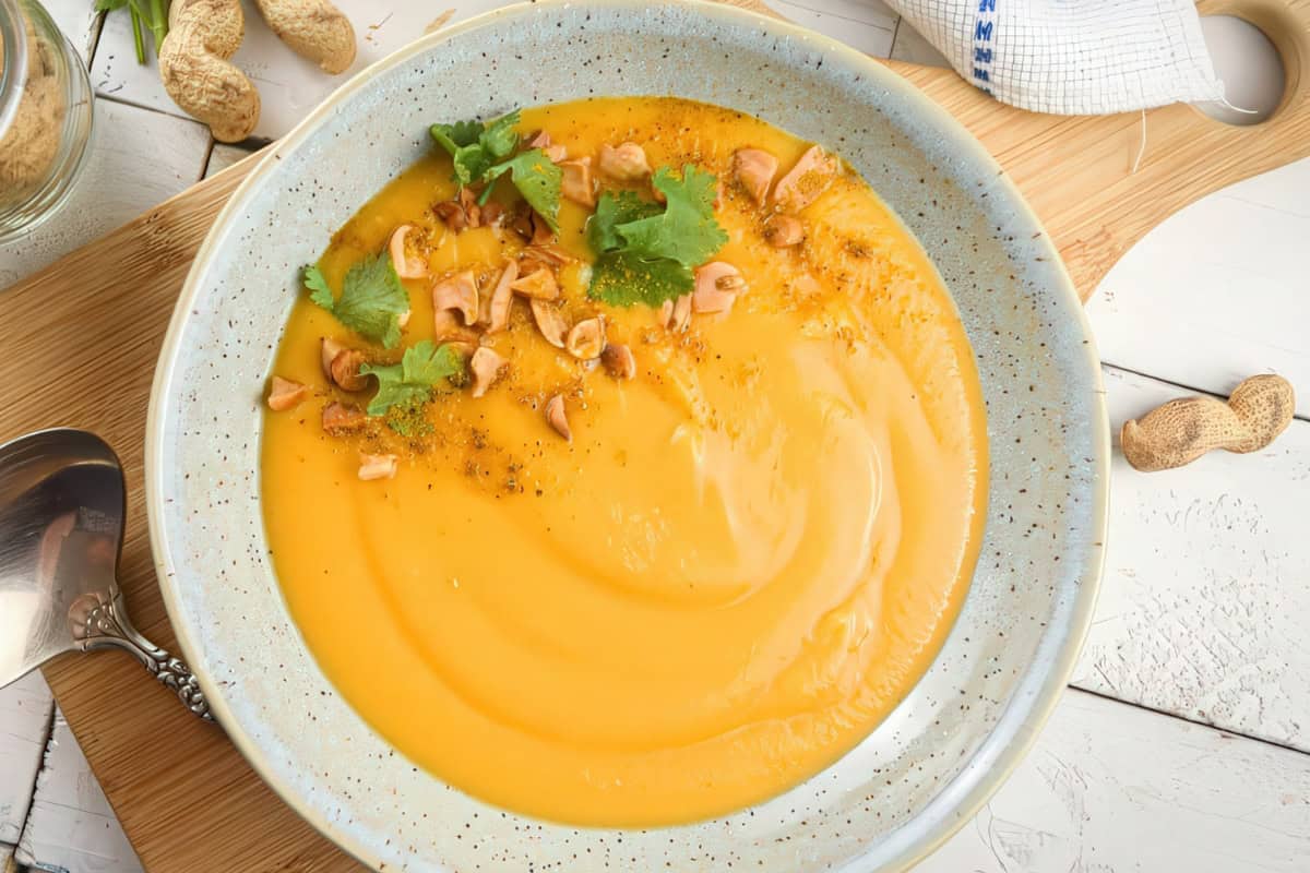A bowl of creamy orange soup garnished with chopped peanuts and fresh cilantro. The bowl is speckled and placed on a wooden board with a spoon nearby. Whole peanuts and a cloth are visible in the background.