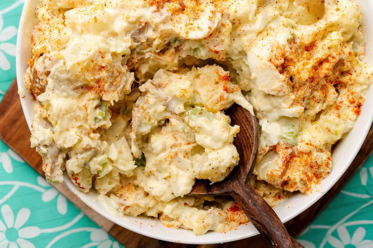 A bowl of potato salad topped with paprika, with a wooden spoon scooping a portion nearby. The salad has visible chunks and rests on a floral-patterned tablecloth.
