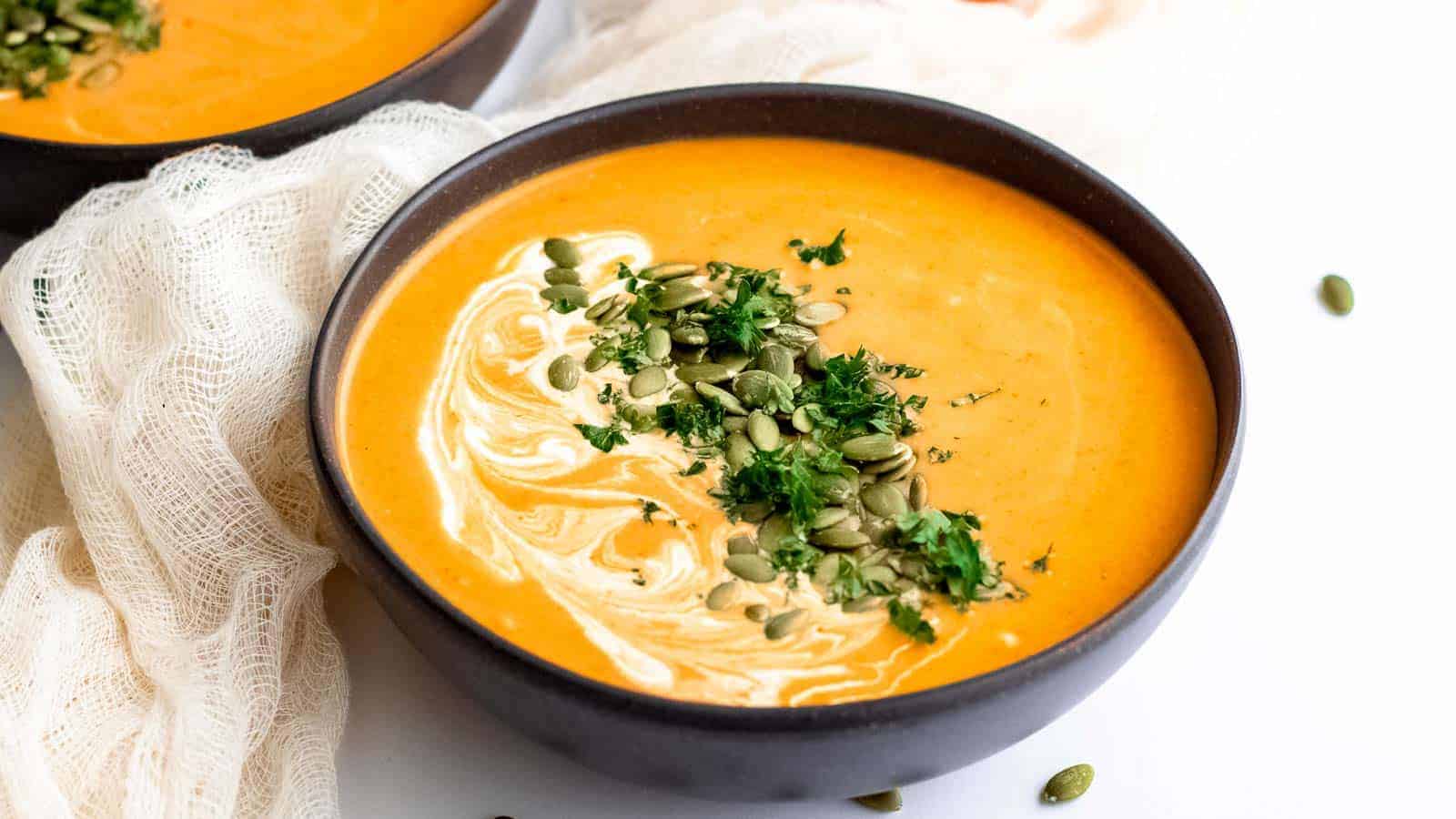 A bowl of orange soup with swirls of cream, pumpkin seeds, and chopped herbs.
