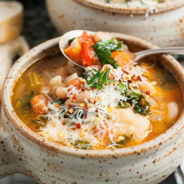 A ceramic bowl of vegetable and white bean soup topped with grated cheese and herbs, with a spoon lifting some and bread behind.