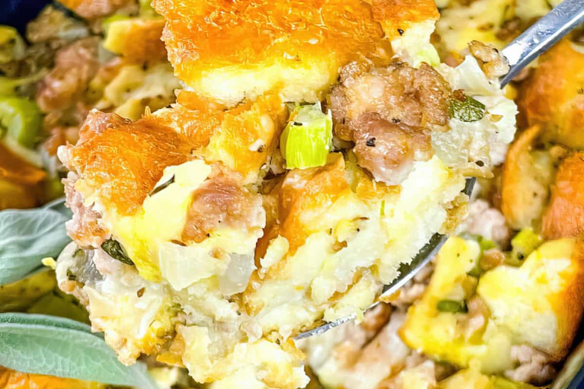 A spoon lifting a portion of stuffing with bread, sausage, celery, and herbs, featuring a crispy top and soft interior.