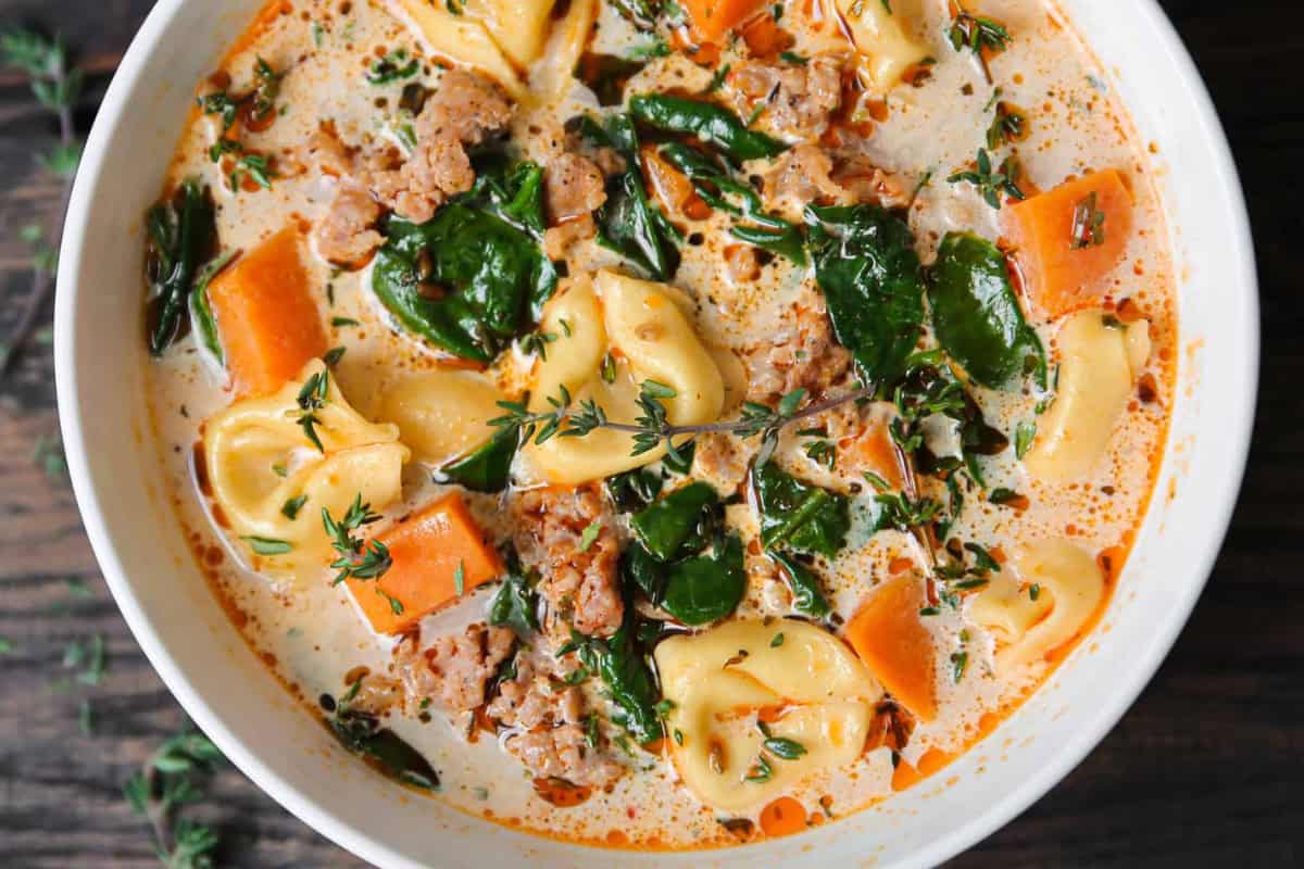 A close-up of a creamy soup featuring tortellini, chunks of sausage, spinach, and carrots, garnished with fresh thyme. The soup is served in a white bowl, showcasing a rich blend of colors and textures.