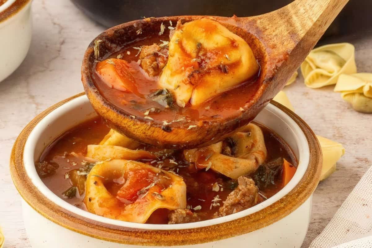A close-up of a wooden spoon holding tortellini over a bowl of hearty tomato soup. The soup is garnished with herbs and contains vegetables, meat, and pasta pieces. A small dish and folded pasta are visible in the background.