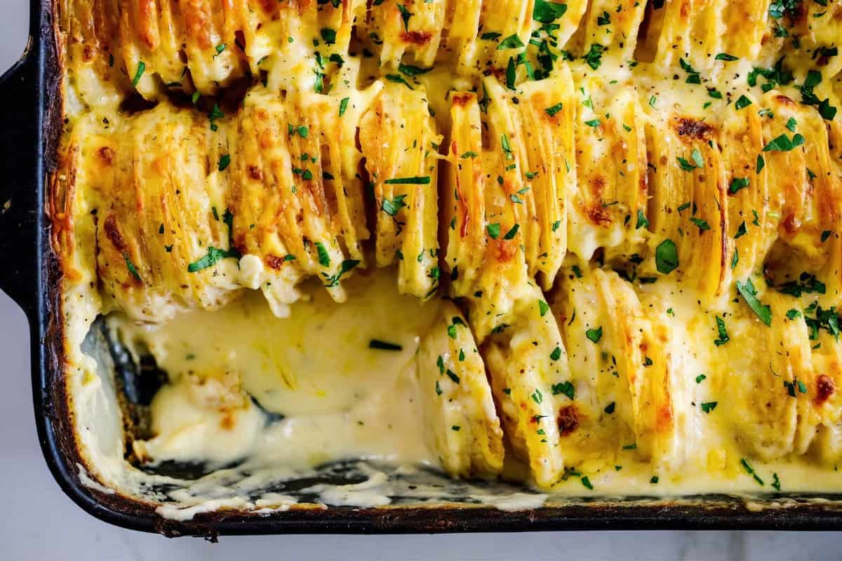 The image shows a baking dish filled with creamy scalloped potatoes topped with chopped parsley. A portion has been removed, revealing layers of sliced potatoes and sauce inside.