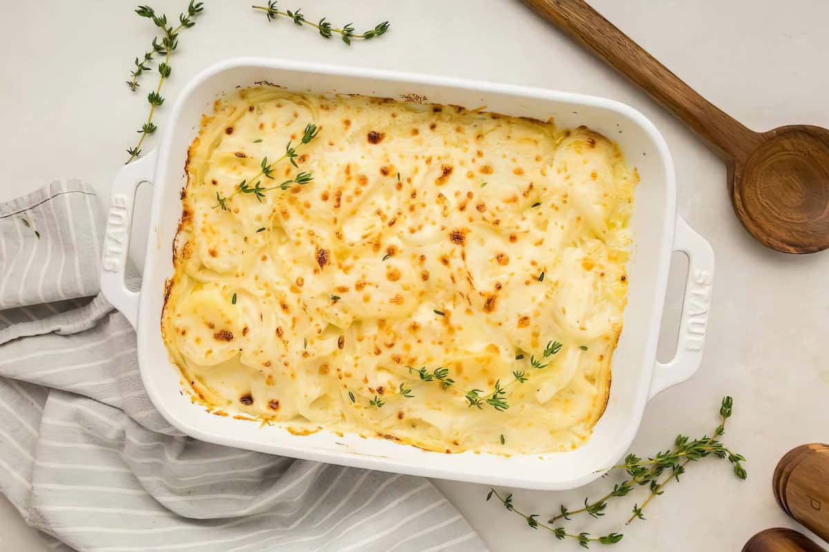 A dish of scalloped potatoes with a golden-brown surface, garnished with fresh thyme sprigs, in a white rectangular baking dish. Nearby are wooden spoons and a cloth.