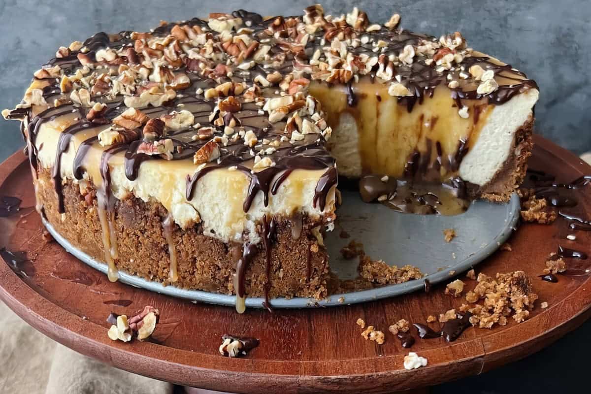 A wooden tray holds a cheesecake topped with chocolate drizzle, caramel sauce, and chopped nuts. A slice is missing from the cake.