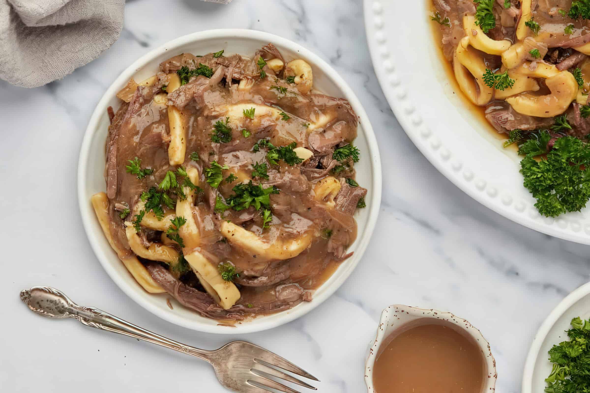 A plate filled with thick egg noodles and tender beef in a rich brown gravy. The dish is garnished with chopped parsley. A small bowl of gravy sits beside the plate, and a silver fork rests nearby on a marble surface.
