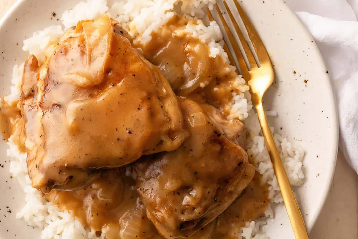 A serving of chicken smothered in brown gravy atop white rice on a plate. A gold fork rests on the side, and a white napkin is partially visible.