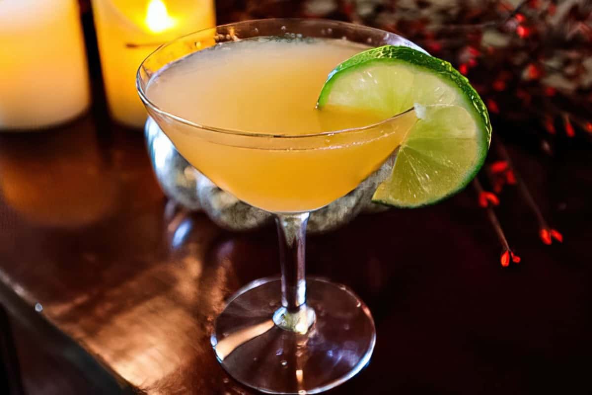 A martini glass containing a cocktail is garnished with a lime slice and placed on a dark surface. A lit candle glows in the background. Multiple other cocktails are arranged nearby.