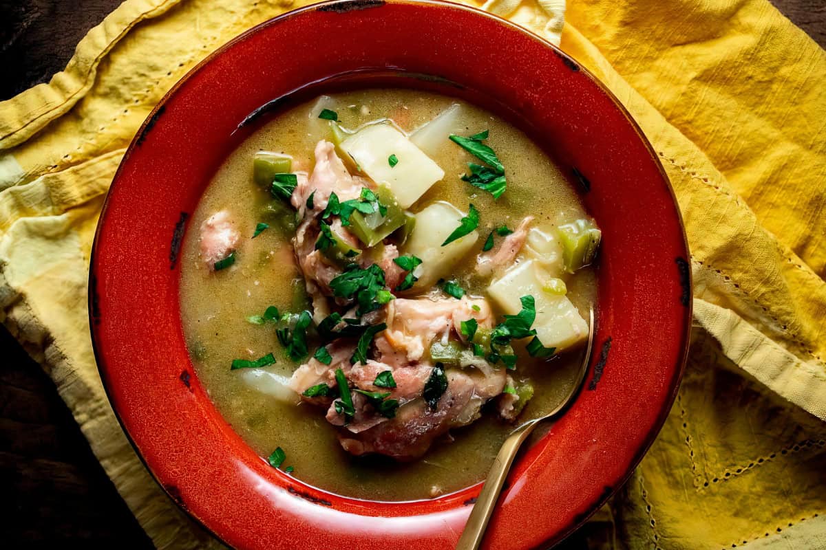 A red bowl filled with soup containing chunks of meat, potatoes, and herbs sits on a yellow cloth.
