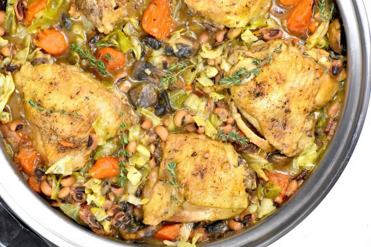 The image features a pot filled with cooked chicken thighs accompanied by carrots, mushrooms, cabbage, black-eyed peas, and herbs.