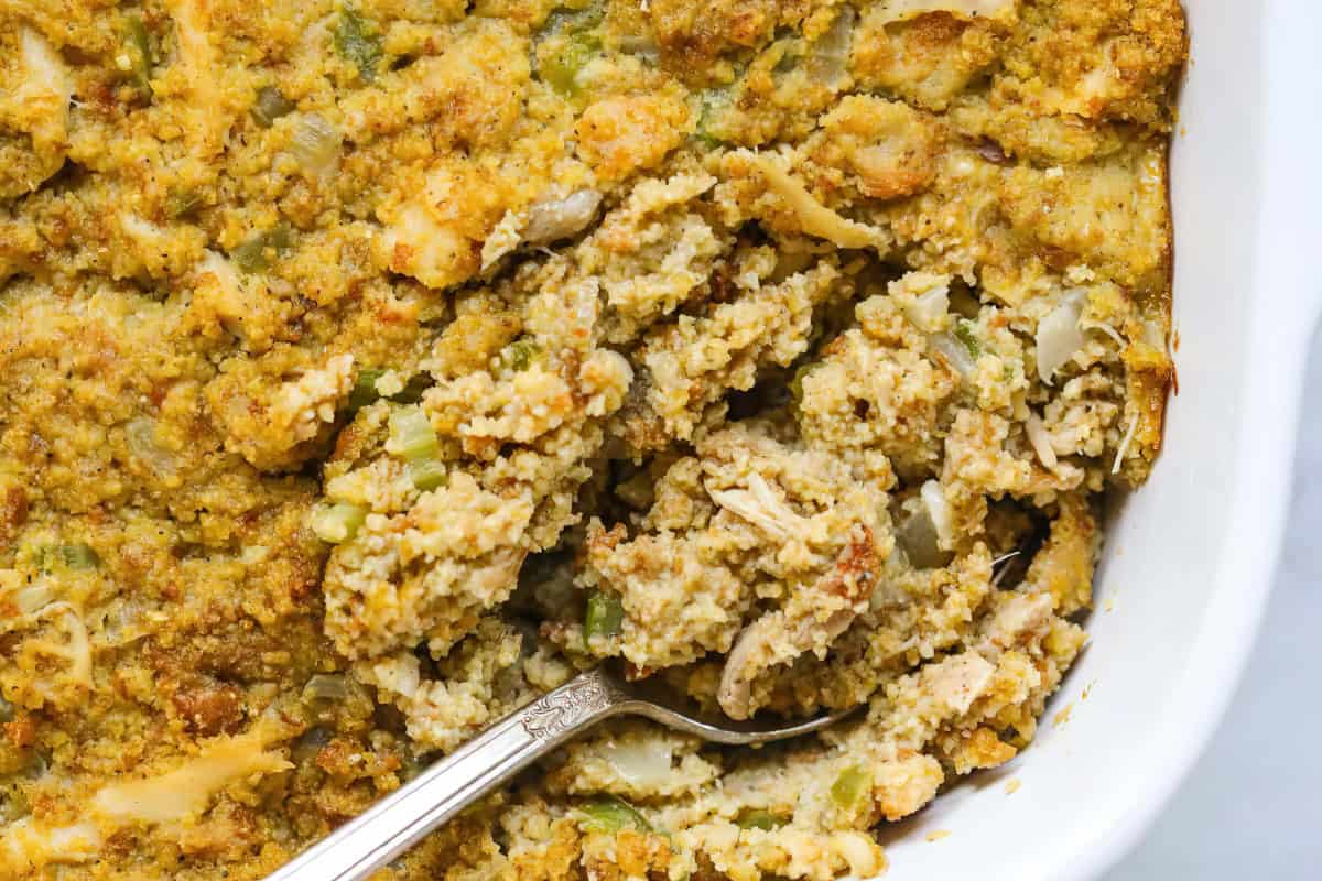 A casserole dish containing stuffing made with bread, celery, and onions, accompanied by a silver spoon.