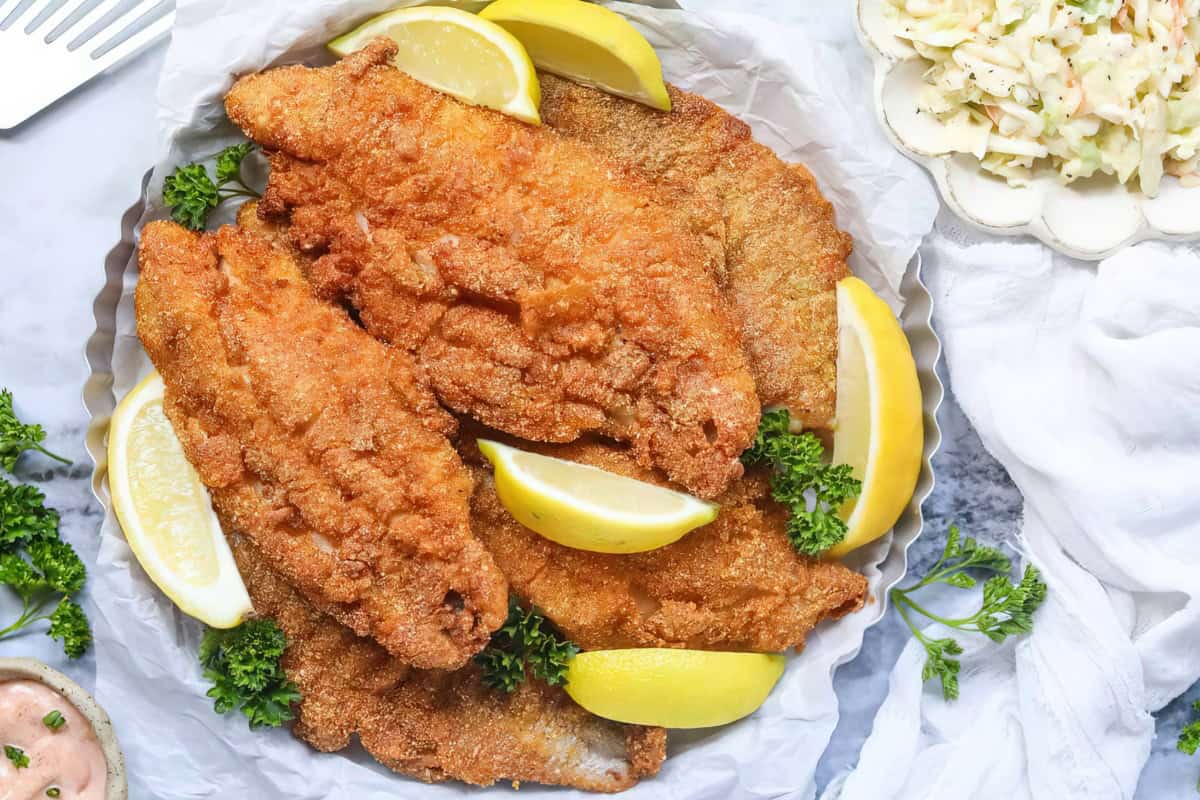 The image features golden fried fish fillets, lemon wedges, parsley, and a side of coleslaw.