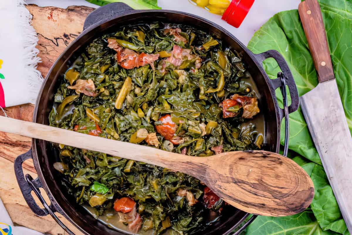 A pot containing collard greens and meat, with a wooden spoon on top of a wooden board.