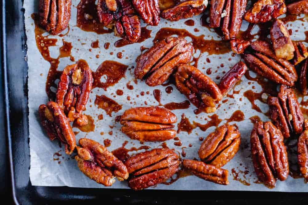 A baking sheet lined with parchment paper holds candied pecans coated in a shiny, caramelized glaze. The nuts are scattered unevenly and glisten under the light.