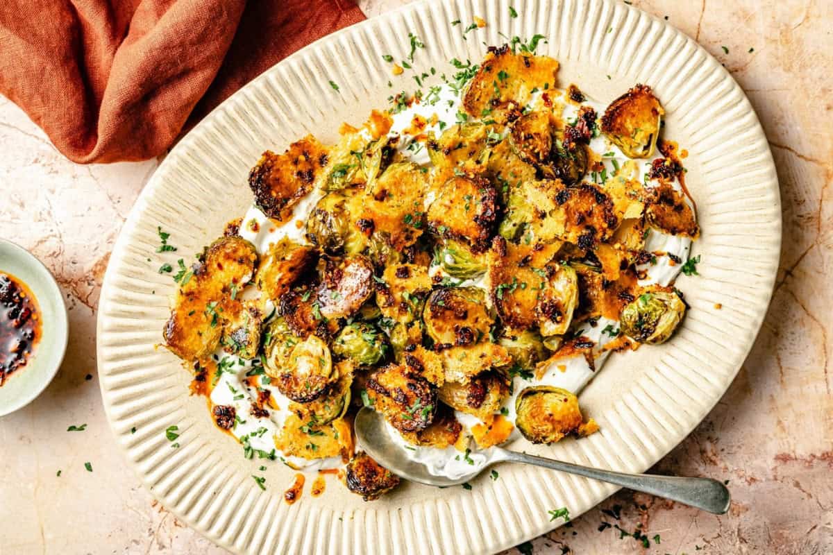 A platter of roasted Brussels sprouts topped with melted cheese and herbs. The dish sits on a textured beige surface with a spoon resting on the platter. A red napkin and a small dish of sauce are visible beside it.