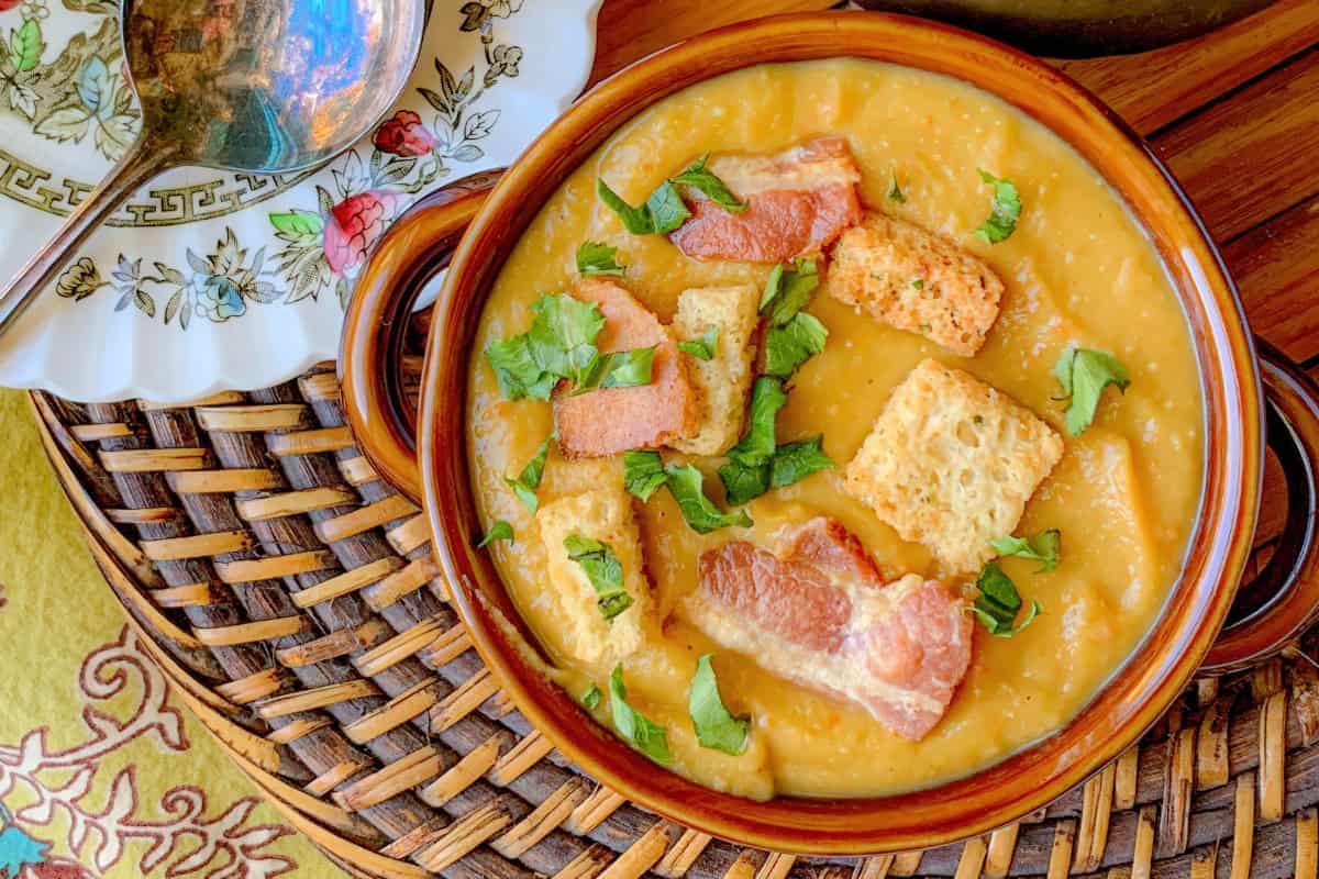 A bowl of creamy soup topped with crispy bacon bits and croutons, garnished with fresh herbs. The soup is served in a brown bowl on a wicker mat, with a decorative saucer and spoon beside it.