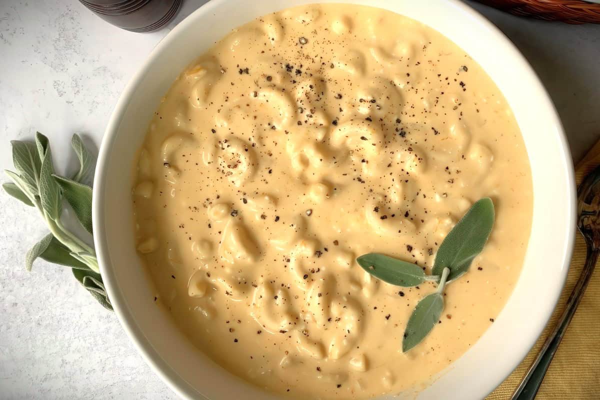 A large bowl of creamy macaroni and cheese garnished with freshly ground black pepper and sage leaves, placed on a white surface.