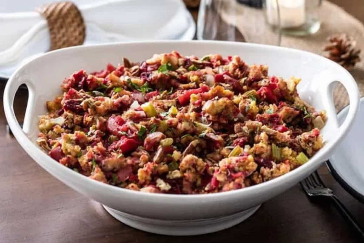 A white serving bowl filled with a colorful mixed salad featuring chopped vegetables, nuts, and herbs. The dish is placed on a wooden table next to plates and a fork, creating a cozy dining setting.