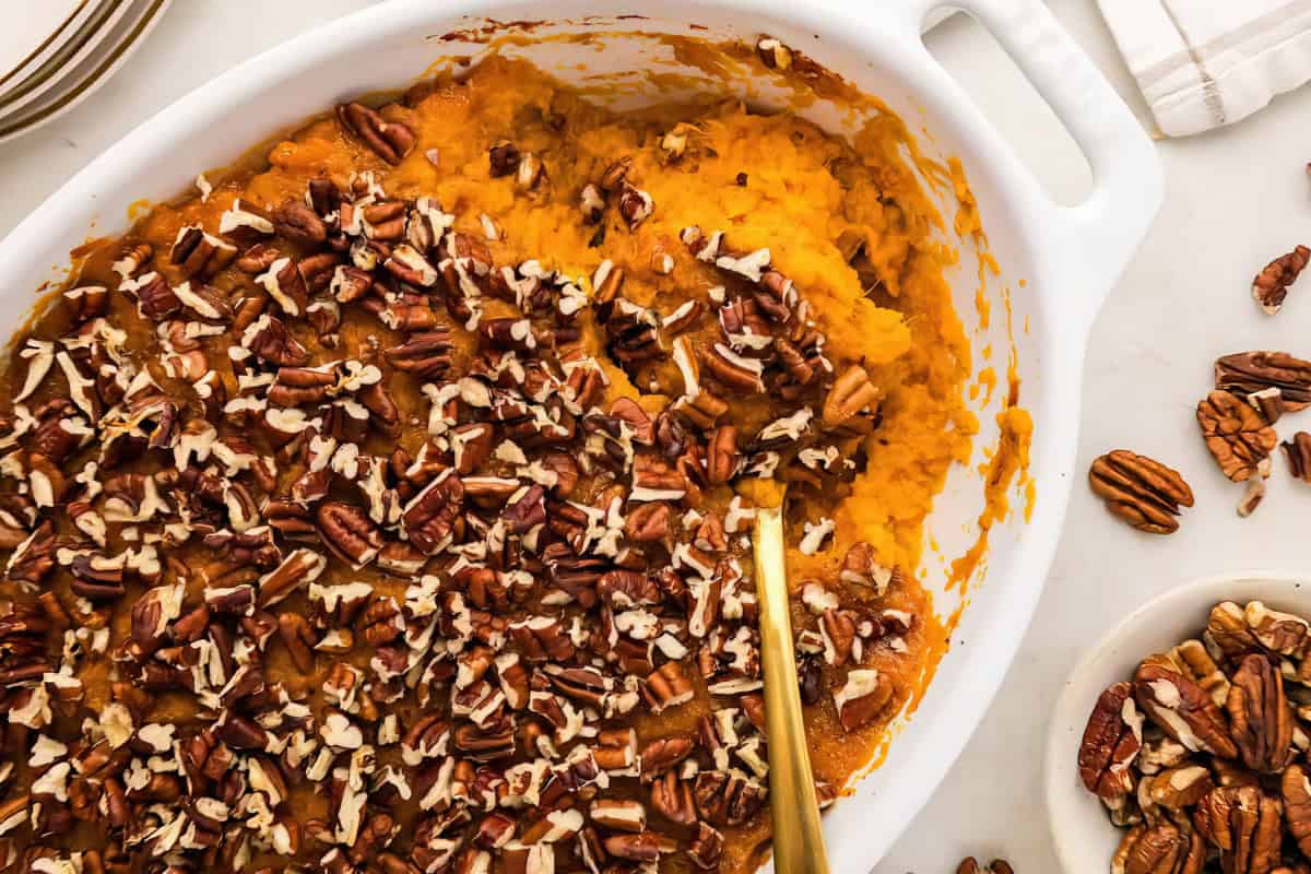 A casserole dish with mashed sweet potatoes, topped with chopped pecans, a serving spoon inside, and extra pecans scattered nearby.