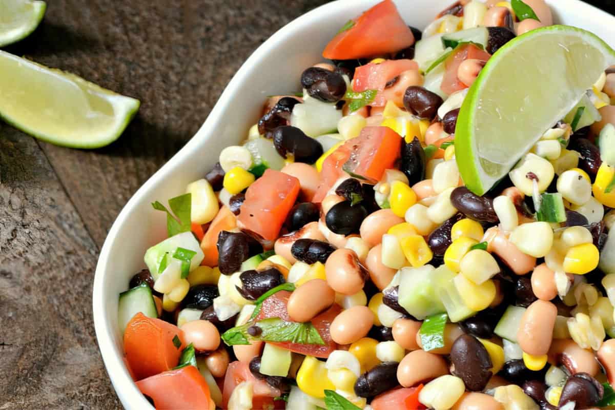 A bowl containing bean salad, corn, tomato, cucumber, and herbs, garnished with a lime wedge.