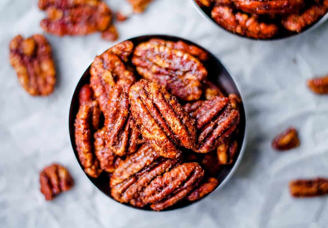 A bowl filled with glazed pecans sits on a light, textured surface. A few pecans are scattered around the bowl, displaying their glossy, caramelized coating. Another bowl with pecans is partially visible in the background.