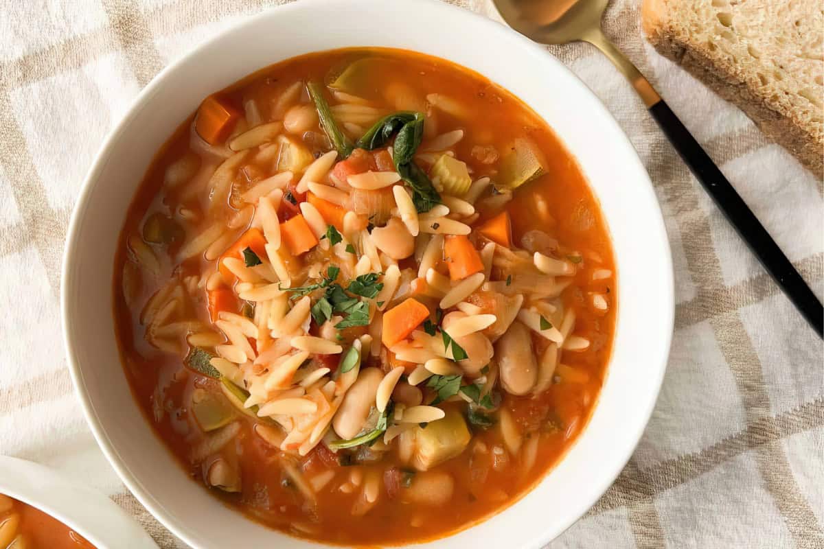A bowl of vegetable orzo soup containing carrots, beans, and greens is placed on a checkered cloth alongside a spoon and a slice of bread.