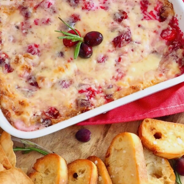 A white casserole dish filled with melted cheese, topped with cranberries and rosemary. It is surrounded by sliced bread and crackers, all placed on a red and white cloth.
