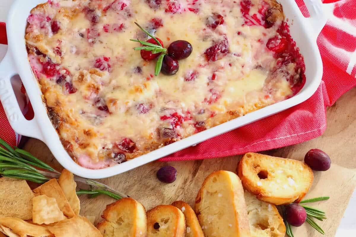 A white casserole dish filled with melted cheese, topped with cranberries and rosemary. It is surrounded by sliced bread and crackers, all placed on a red and white cloth.