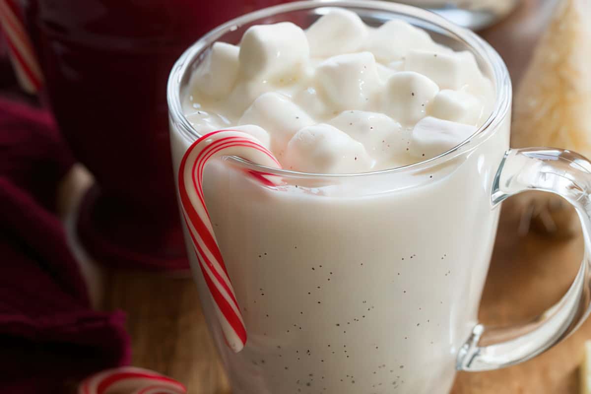 A clear glass mug filled with creamy eggnog topped with marshmallows. A candy cane hangs on the rim, adding a festive touch. The drink is on a wooden table, with part of a red cloth visible in the background.